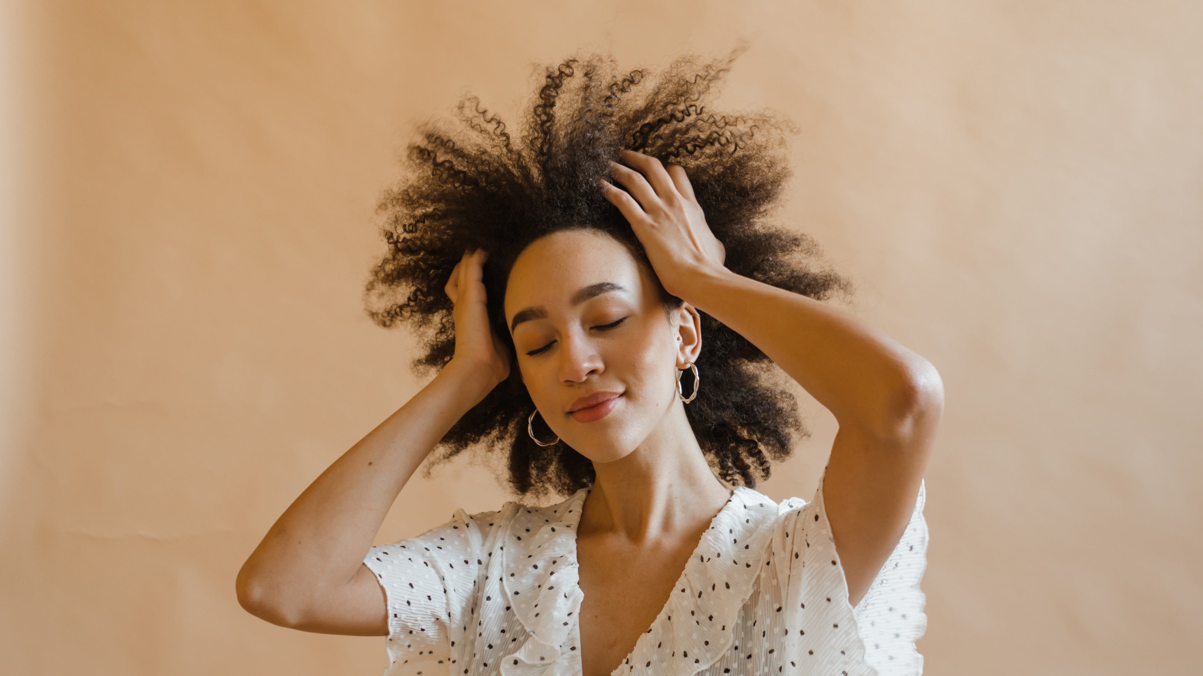 Woman with curly hair wearing a white polka dot dress against a beige background